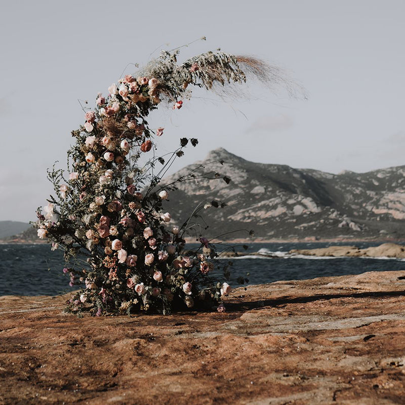 Stunning curved floral arrangement with neutral coloured roses and foliage sits on a rocky outcrop in front of the ocean, there is a mountain behind