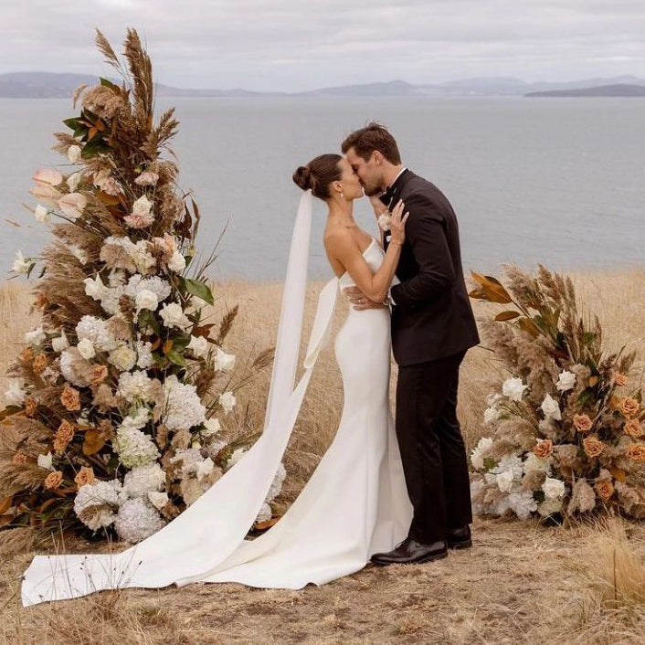Photograph of a married couple kissing between two stunning stands of roses in muted nude colours, the ocean is behind them