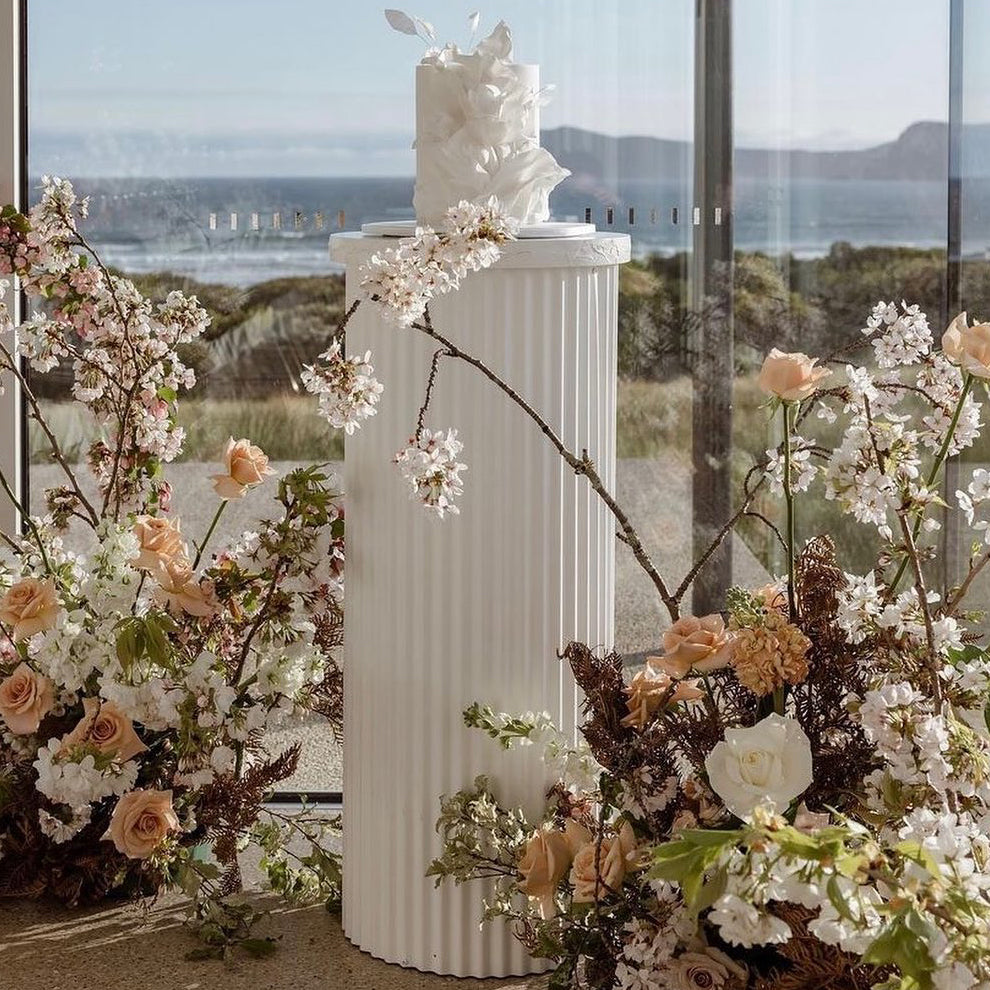 Photograph of a white column with a cake on top, it is surrounded by stunning flowers