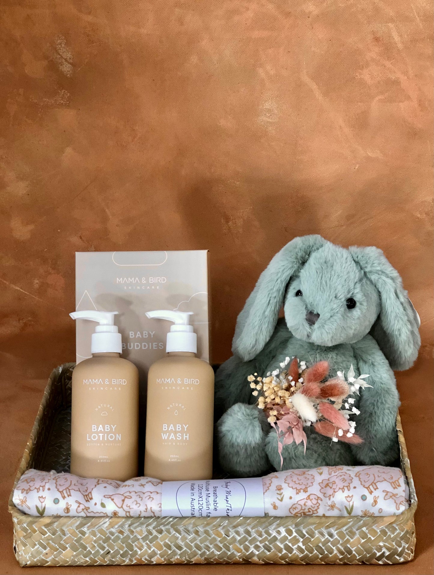 Photograph of a basked containing a beautiful floral baby muslin, luxe baby lotion and wash and a fluffy blue rabbit holding some dried flowers