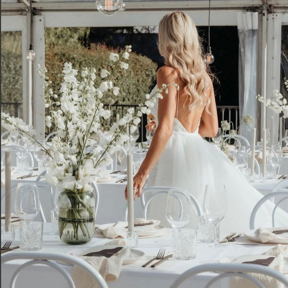 Photograph of a bride in an etherial white gown walking past beautiful white reception tables with sprays of white flowers