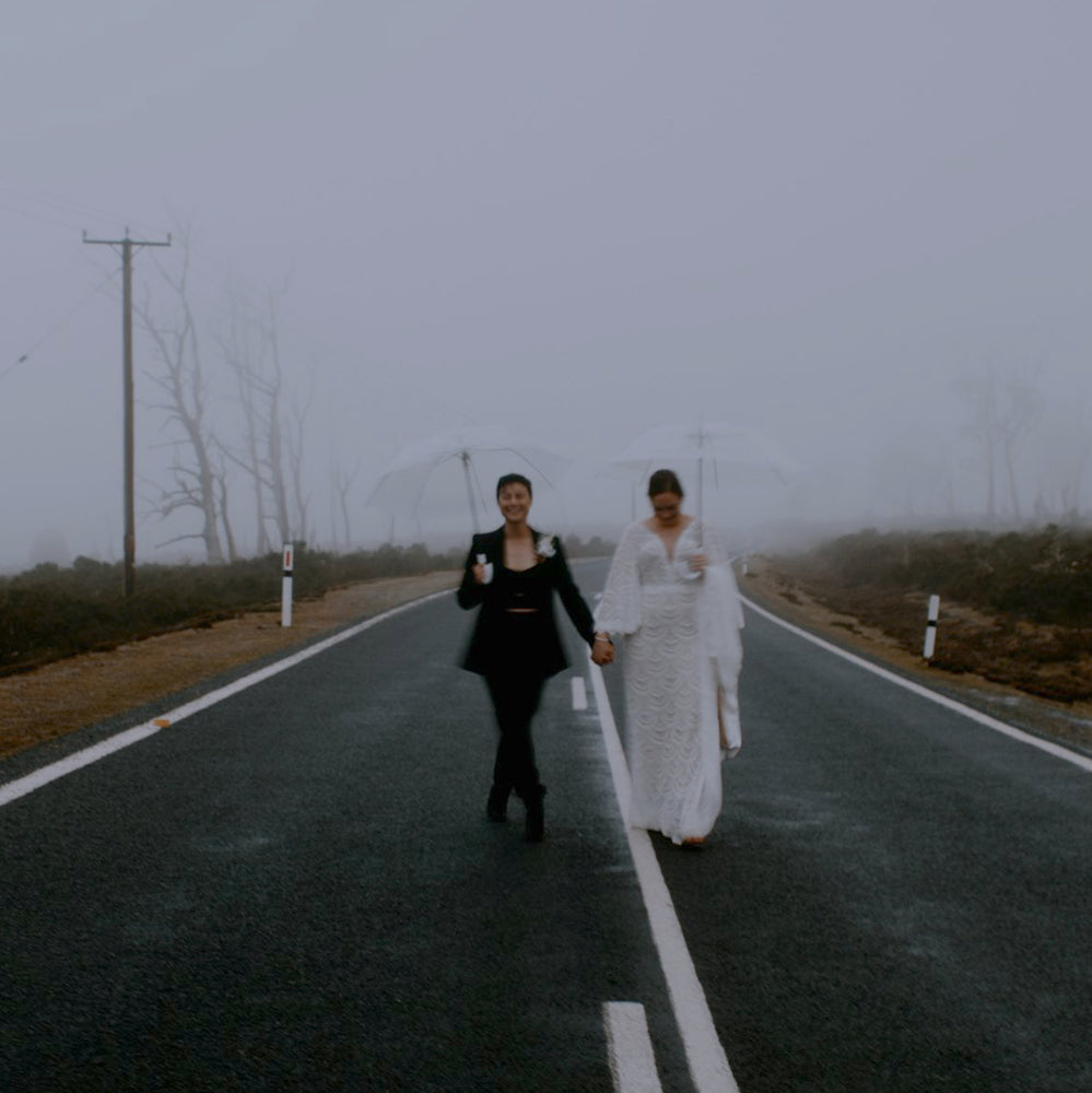 Two married women stand hand in hand in the middle of a country road in the mist