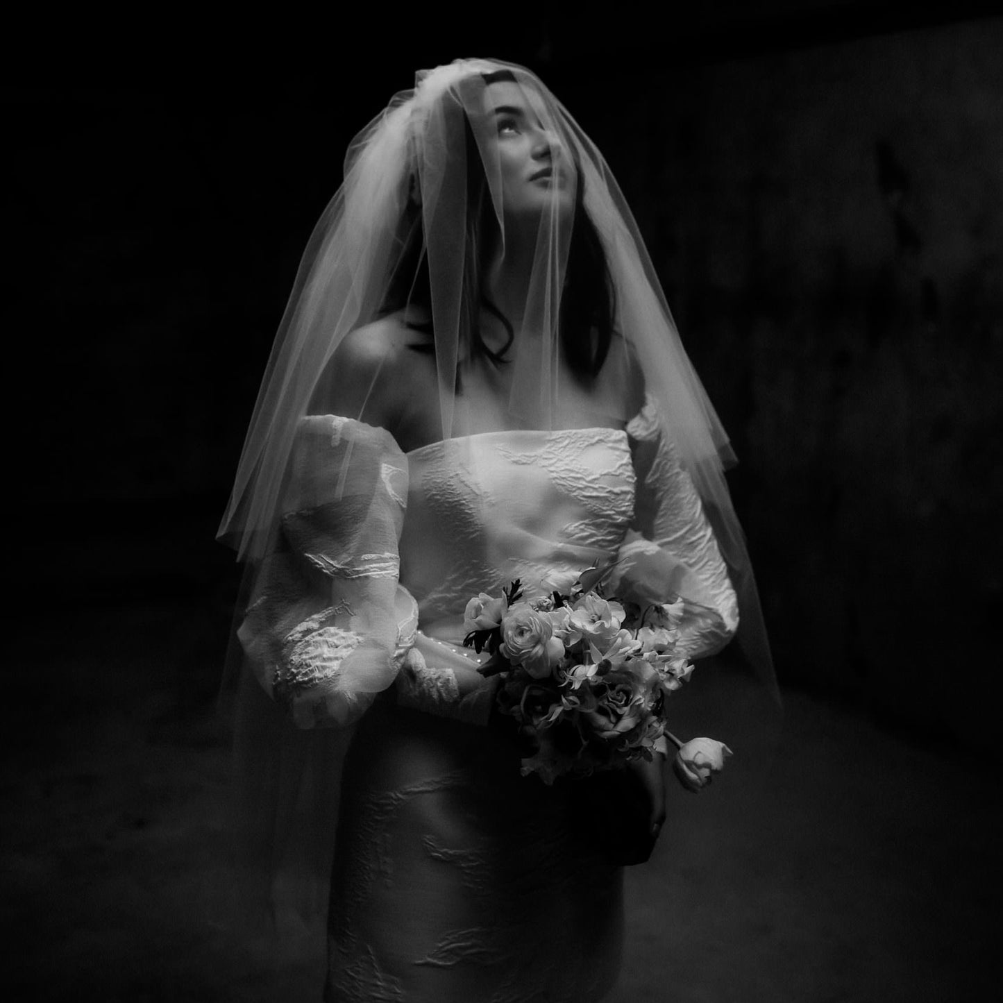 Striking photograph in black and white of a dark haired young woman wearing a veil and wedding dress and looking upwards