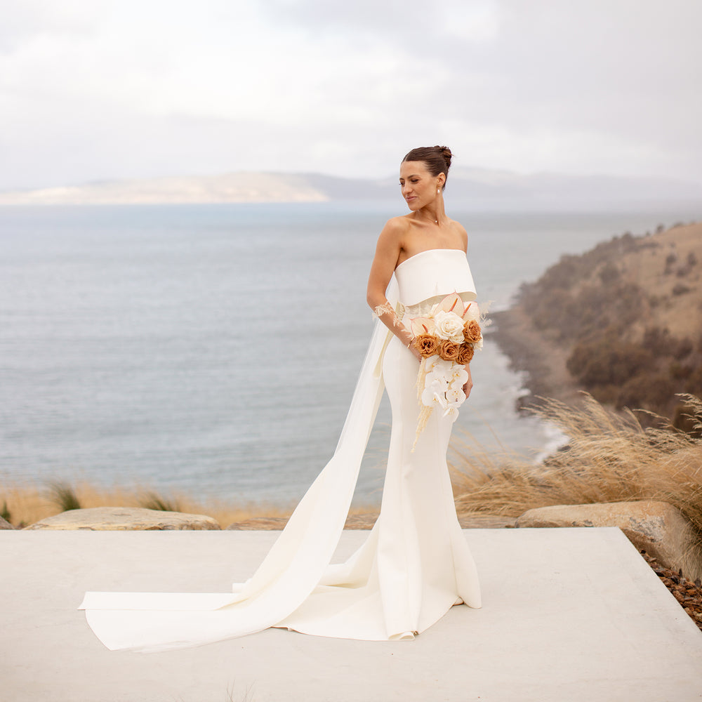 Stunning photograph of a woman wearing an elegant wedding dress, she has nude and white roses in her hands, behind her is the ocean 