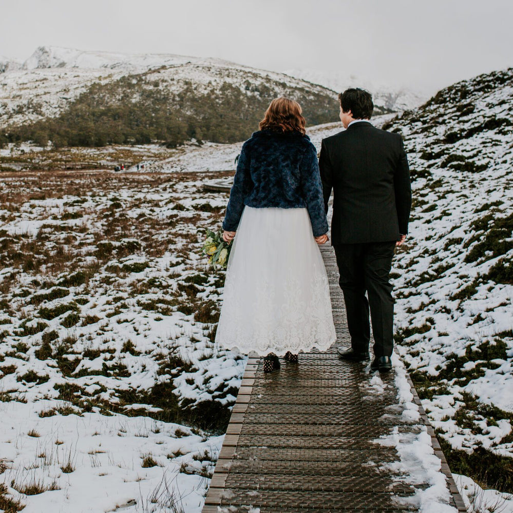A bride and groom walk away from the camera hand in hand, along the boardwalk at Cradle Mountain, there is snow on the ground