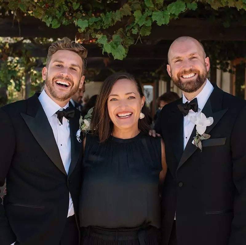 Picture of a dark haired woman standing and smiling between two handsome grooms
