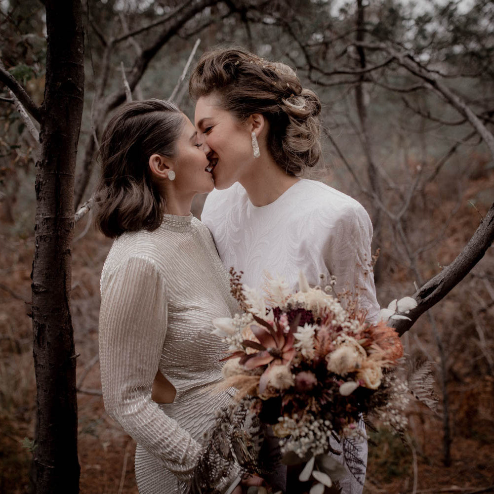 Two women in wedding dresses share a kiss, one is holding a beautiful arrangement of native flowers