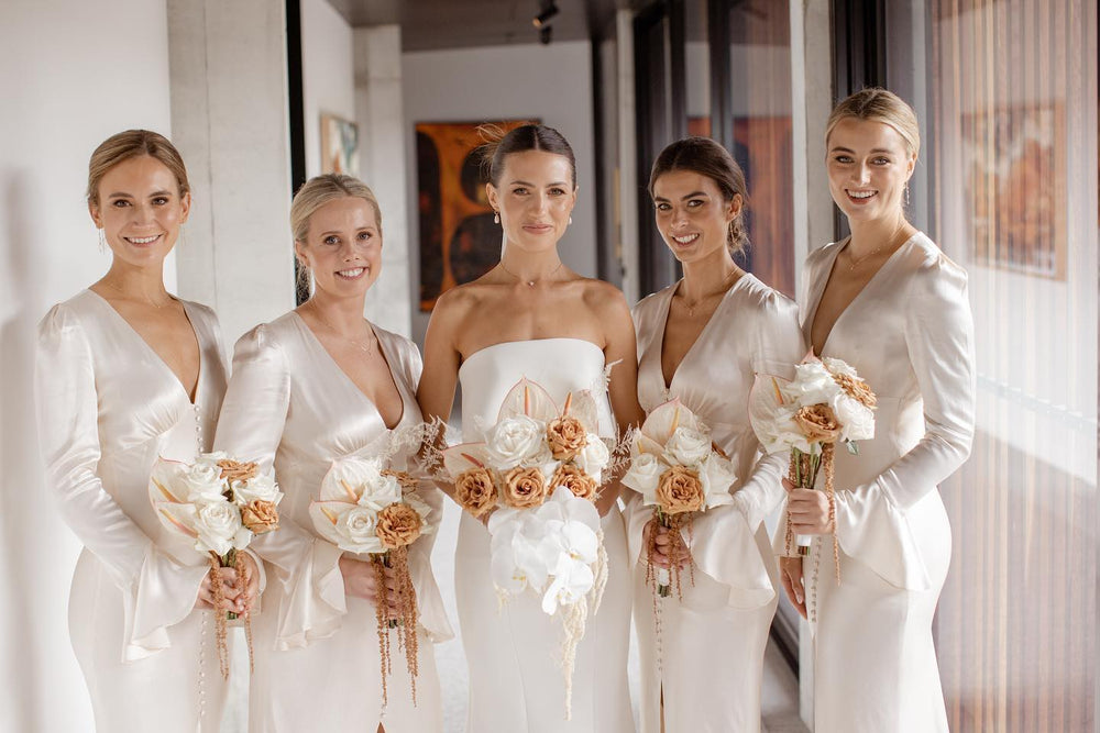 Photograph of a smiling bride surrounded by her brides maids either side, they are wearing white and holding beautiful bouquets in white and neutral colours