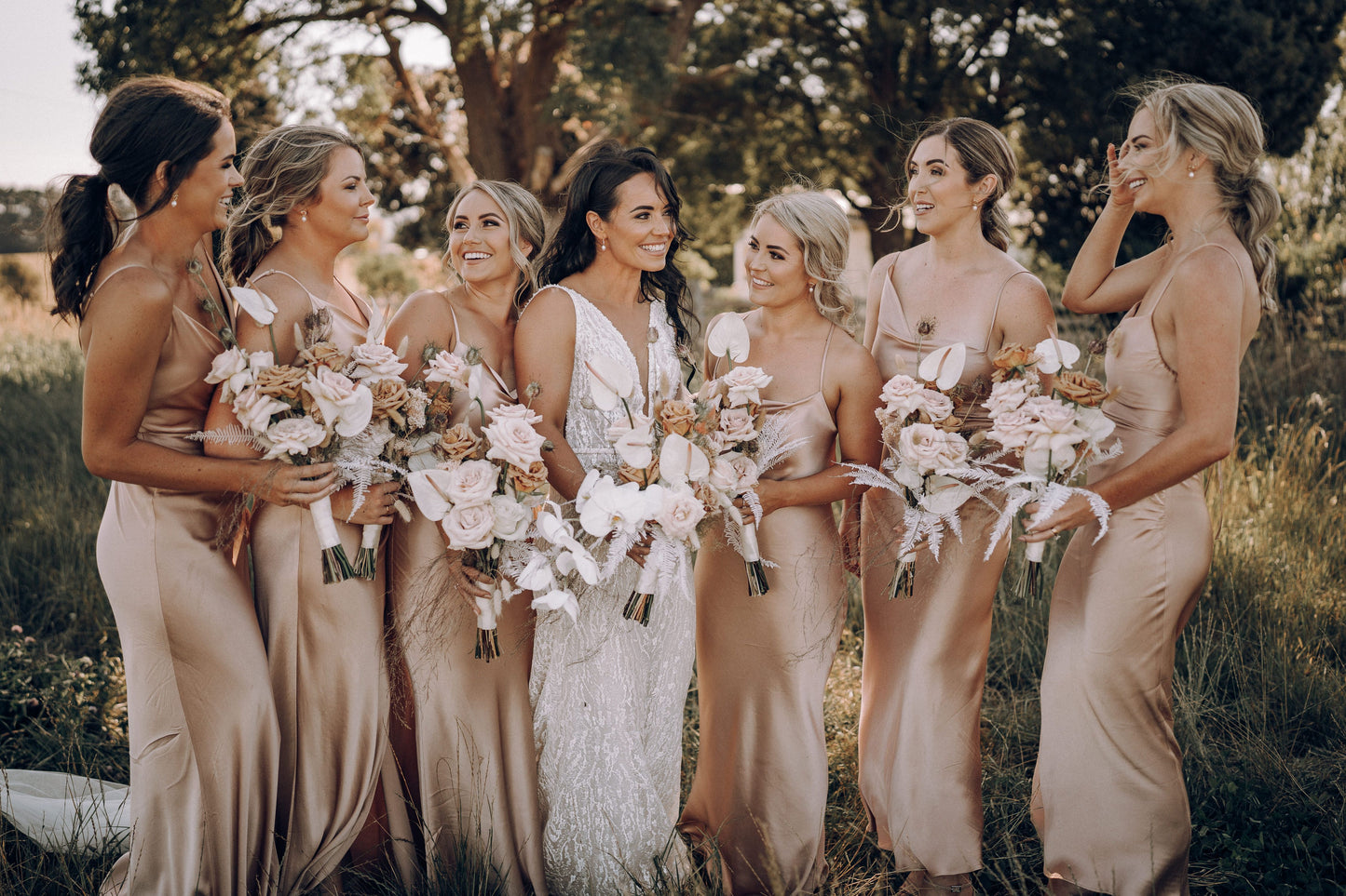 Photograph of smiling brides maids with the bride in white in the middle, they are holding lavish flowers in soft pink and neutral tones